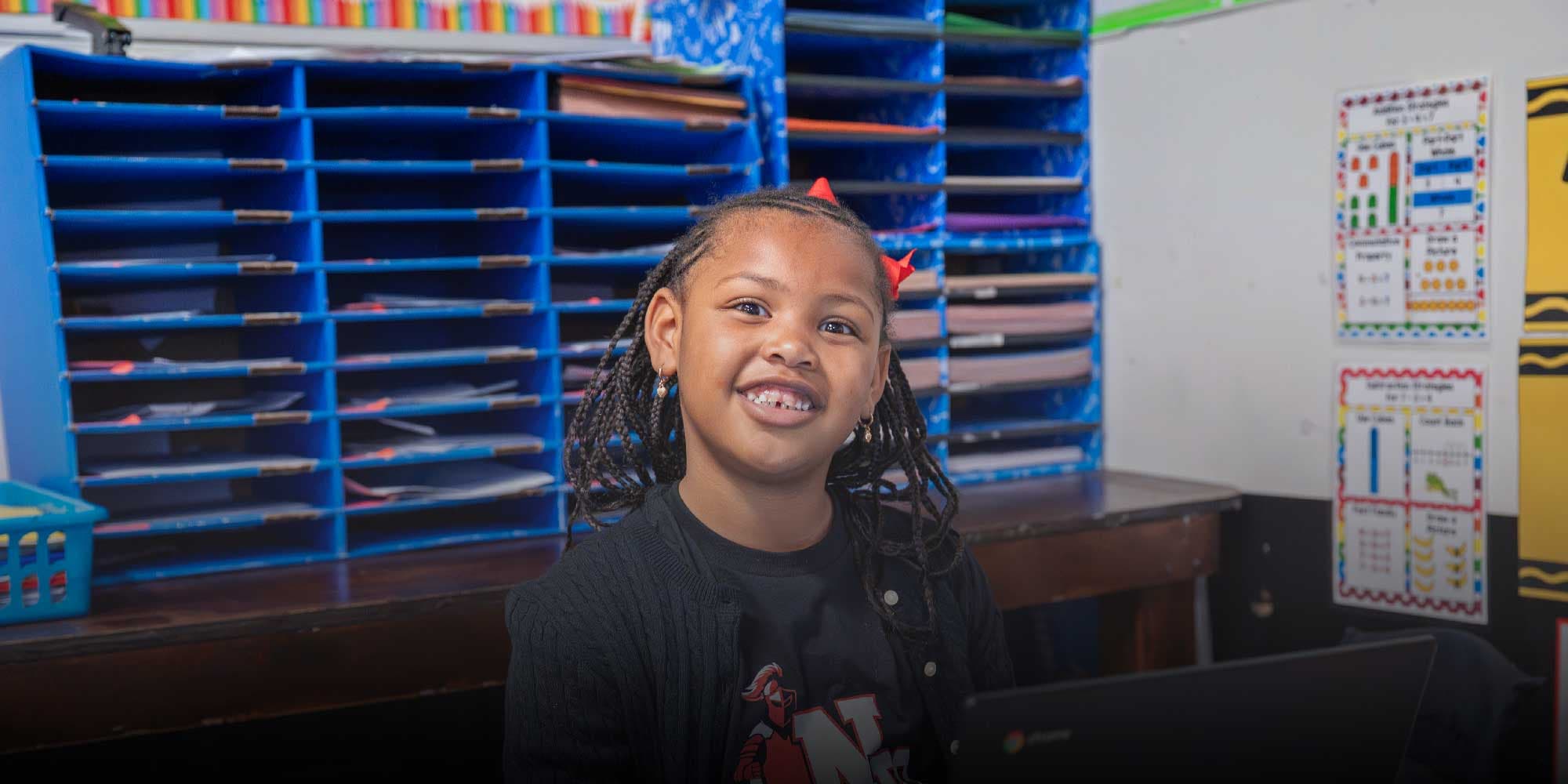 Smiling student at a desk in a classroom with blue cubbies.