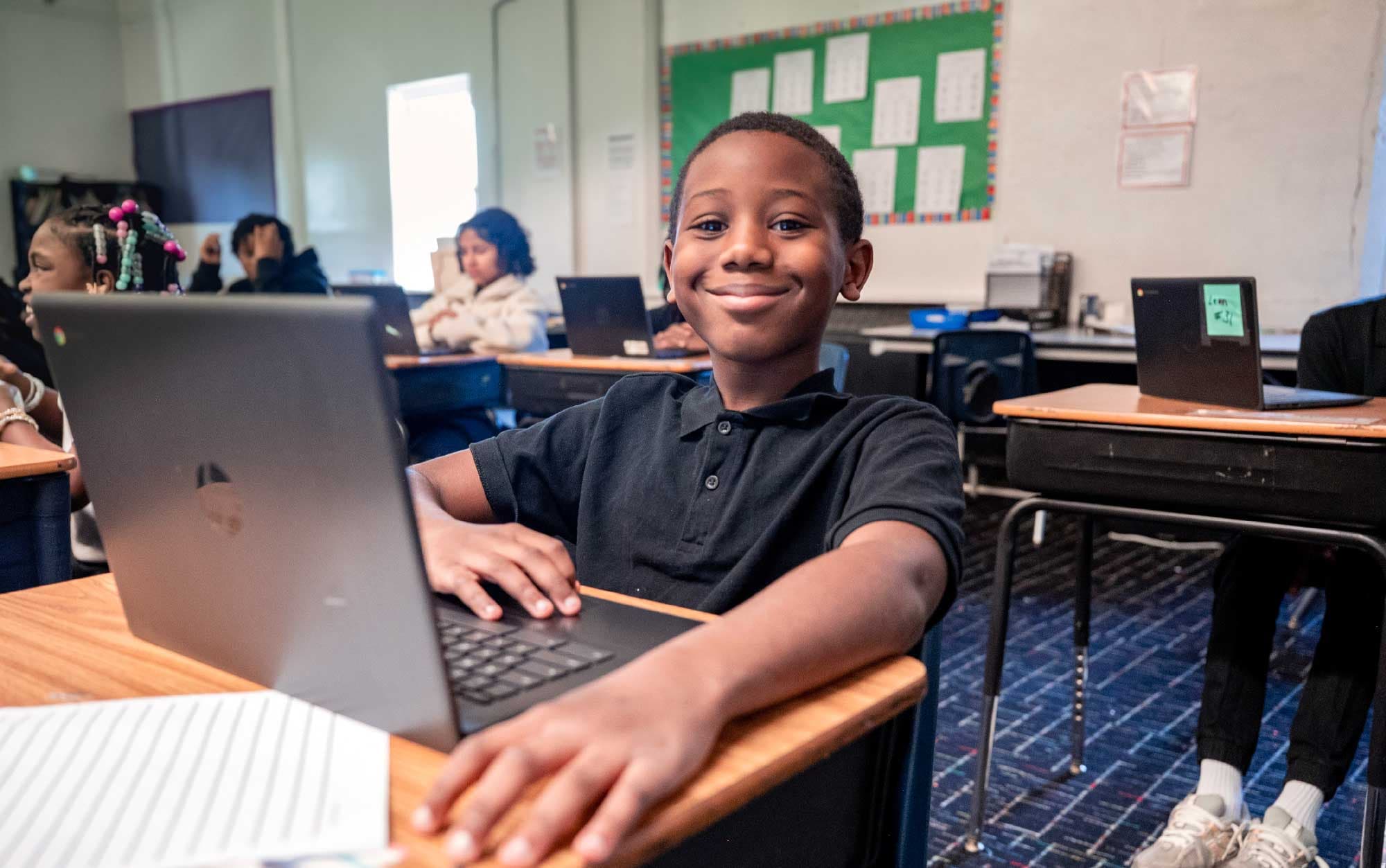 Smiling student in a black polo at a desk with an open laptop.