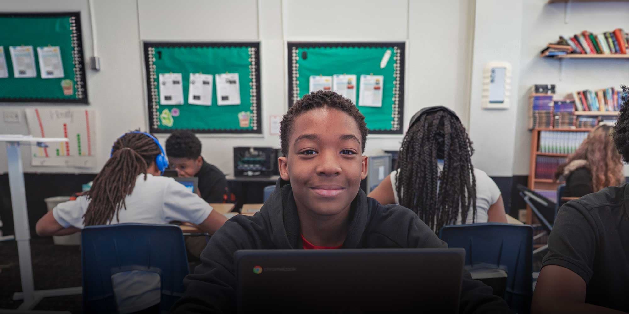 Smiling student at a desk with a laptop in a classroom.