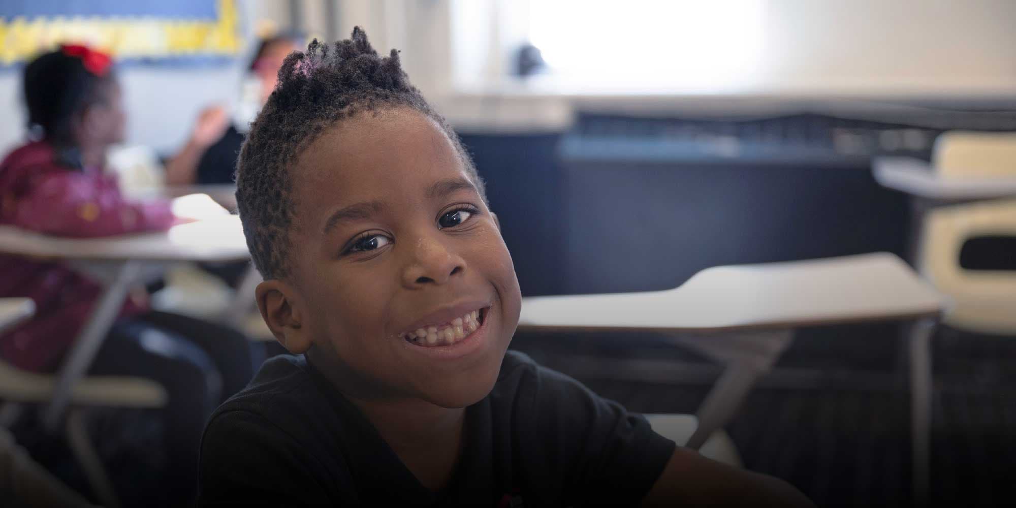 Young student smiling broadly at a classroom desk.