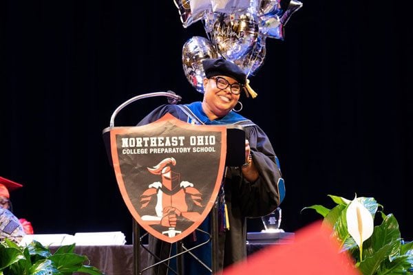 Adult in academic regalia smiles behind a podium with a school logo.