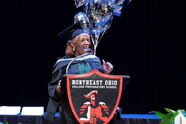 Adult in graduation attire speaking at a podium with balloons.