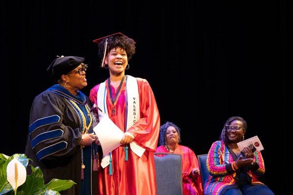 Adult in academic regalia presents document to smiling young adult in graduation gown.