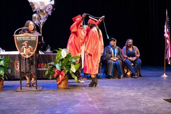 Two adults in red gowns on stage, one turning the other's graduation cap tassel.