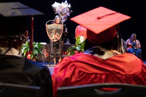 Adult speaking at podium during graduation, viewed from behind two graduates.