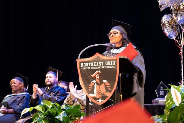 Adult speaking at podium during graduation, others seated and clapping.