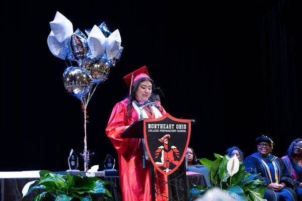 Student in red cap and gown speaking at a podium during graduation.